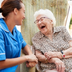 Senior Woman Sitting In Chair And Laughing With Nurse In Retirement Home